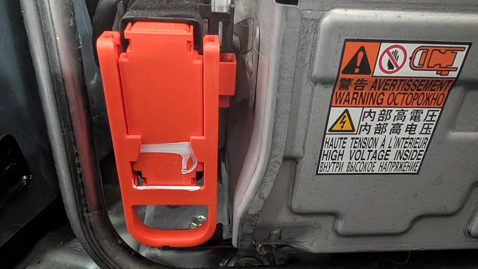 orange high-voltage service plug grip in place on the traction battery, with "HIGH VOLTAGE INSIDE" warning label visible